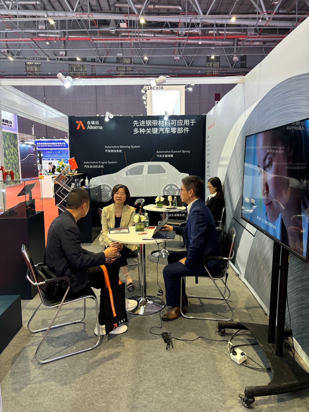 Three business professionals sit around a small round table having a discussion at the Alleima (Shanghai) Materials Technology Co., Ltd. booth during Auto Shanghai 2025. The booth backdrop showcases automotive component applications, including engine systems and steering systems. A fourth person is visible working near a monitor displaying promotional visuals