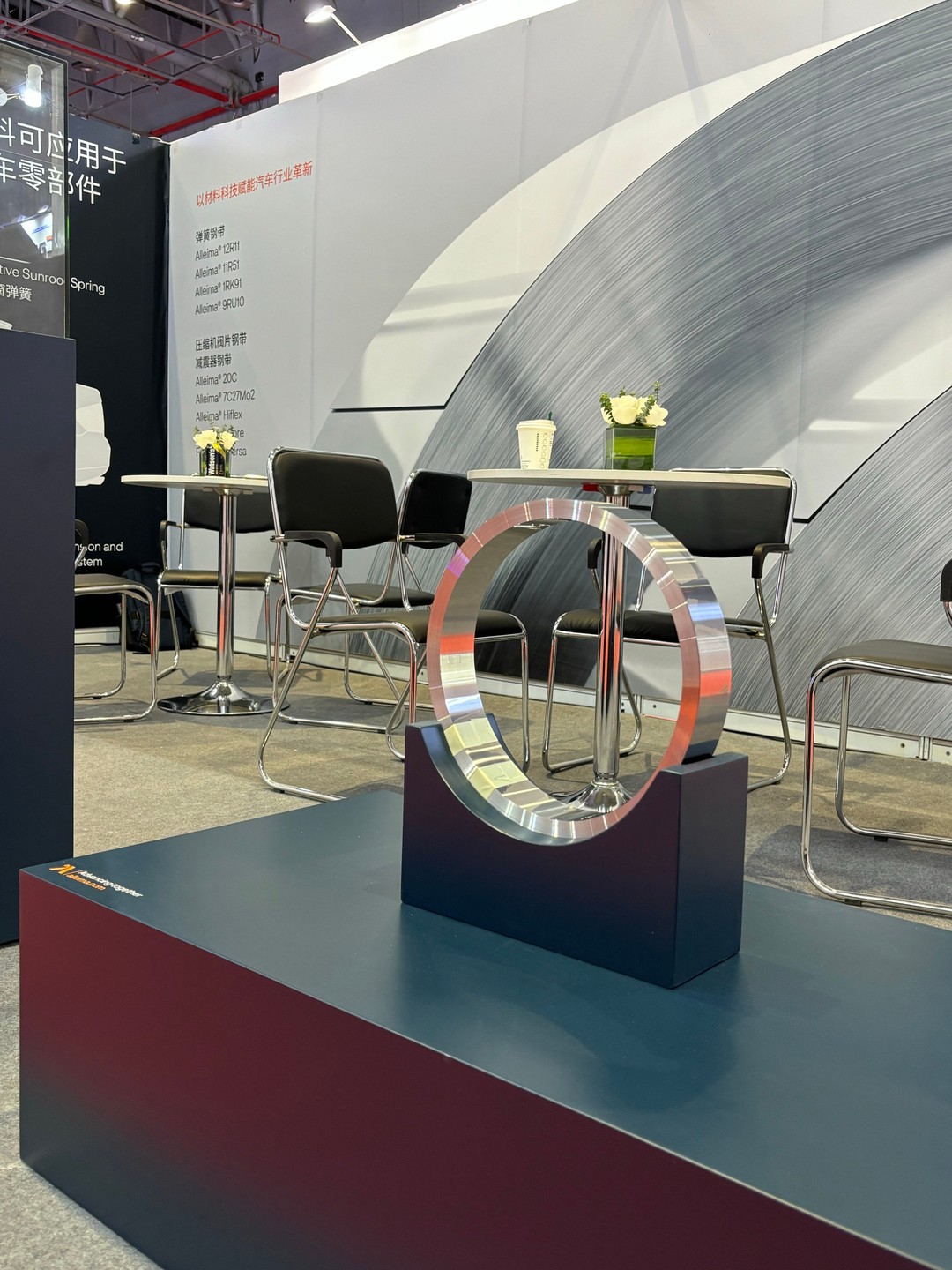 A close-up of a polished metal ring component displayed on a raised pedestal at the Alleima (Shanghai) Materials Technology Co., Ltd. booth during Auto Shanghai 2025. Behind the display is a seating area with chairs and a table, decorated with a small flower arrangement and a coffee cup. The background features a technical illustration and product names in Chinese.