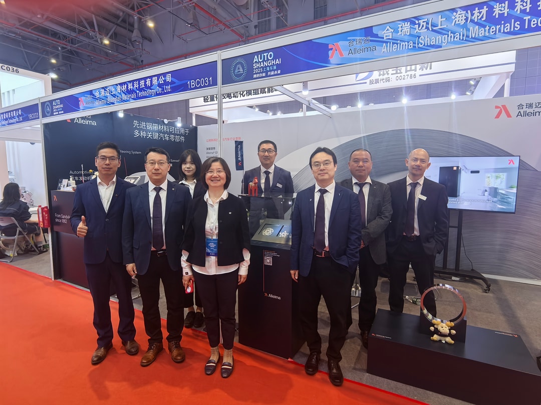 A group of eight professionally dressed representatives from Alleima (Shanghai) Materials Technology Co., Ltd. pose together at their exhibition booth during Auto Shanghai 2025. The booth features branded displays, informational materials, and product samples, with clear signage and a TV screen in the background.