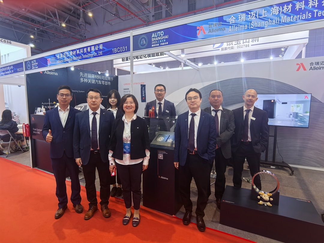 A group of eight professionally dressed representatives from Alleima (Shanghai) Materials Technology Co., Ltd. pose together at their exhibition booth during Auto Shanghai 2025. The booth features branded displays, informational materials, and product samples, with clear signage and a TV screen in the background.